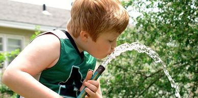 Young boy drinking water from a garden hose outside.