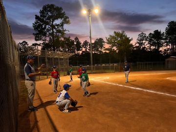 Youth baseball game at sunset under field lights.