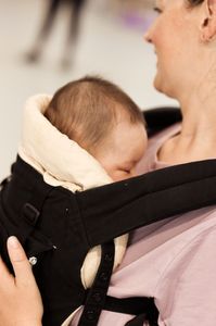 A dark haired baby sleeping in a baby carrier being carried by her Mum. 