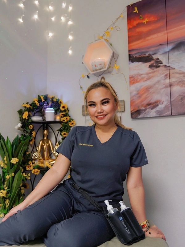Smiling massage therapist in uniform sitting in a cozy, decorated room.