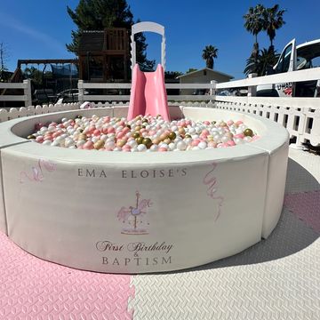 A pink and white ball pit with a slide for a child's birthday and baptism celebration.