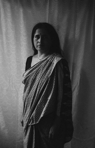 Black and white portrait of a woman in a patterned saree against a textured curtain.