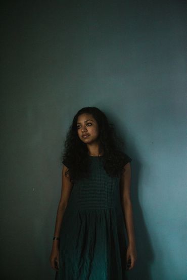 Young woman with curly hair stands against a dark teal wall, deep in thought.
