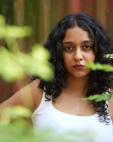 Young woman with curly hair gazes thoughtfully through greenery.