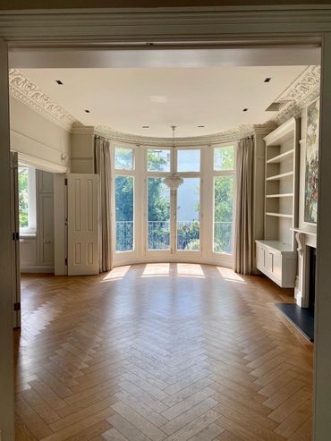 Bright empty room with large bay windows and wooden herringbone floor.