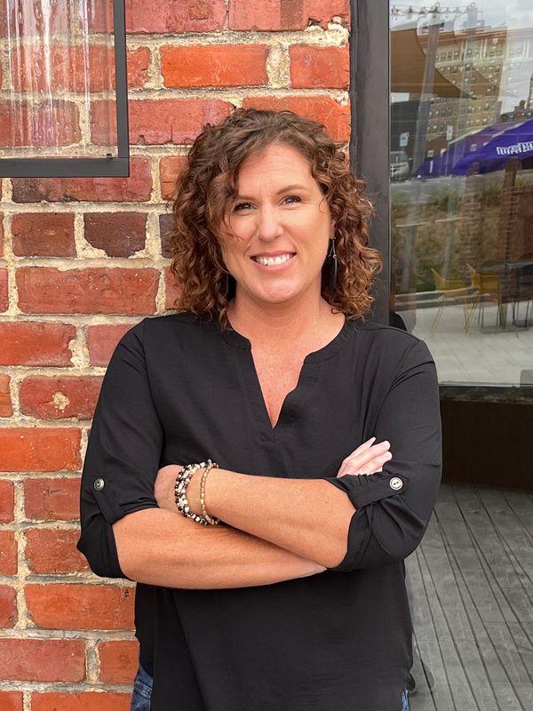 Woman with curly hair smiling, standing with arms crossed against a brick wall.
