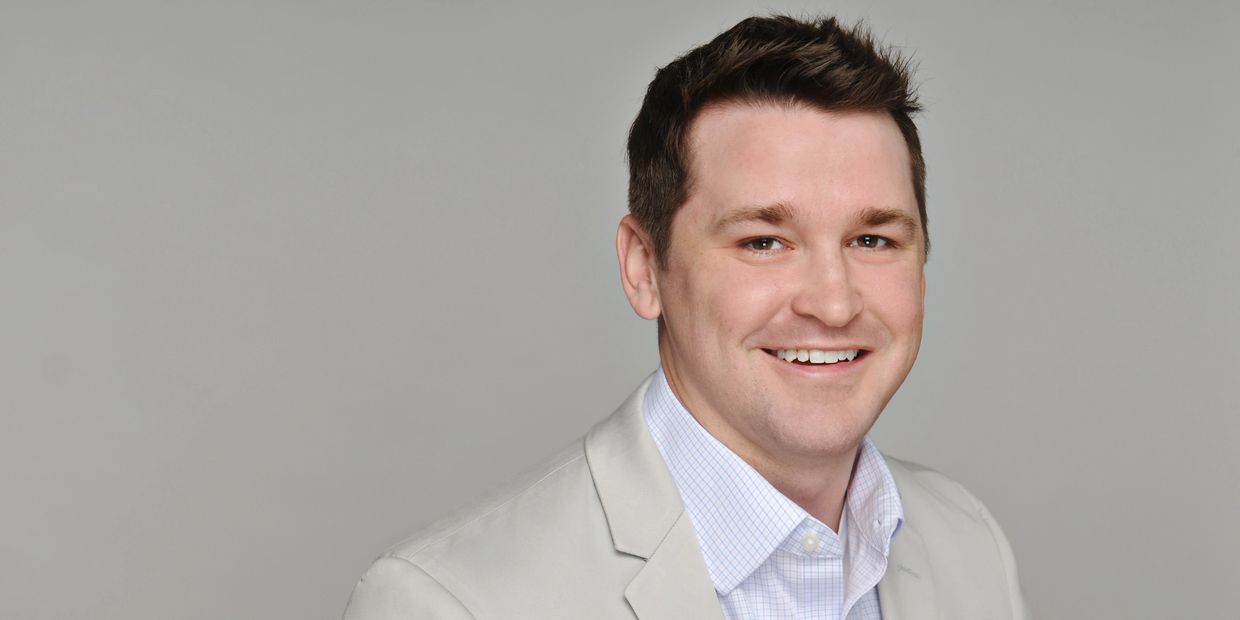 Smiling man in light gray blazer and checkered shirt against plain background.