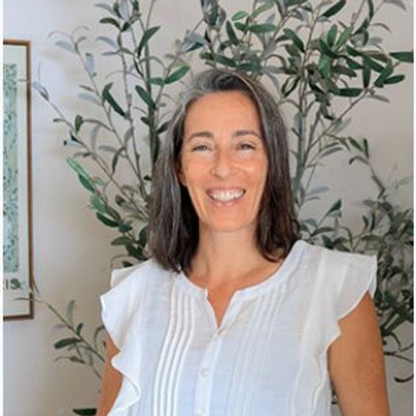 Smiling woman in a white blouse stands in front of green plants indoors.