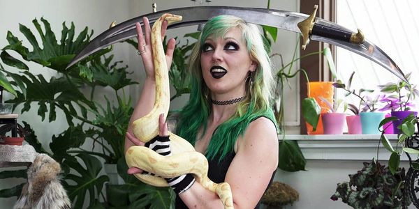 A woman with green hair holds a yellow snake and a large curved sword.