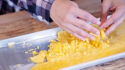 Hands breaking up yellow wax or cheese chunks on a metal tray.