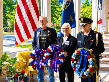 Three people holding patriotic wreaths near American flags outdoors.