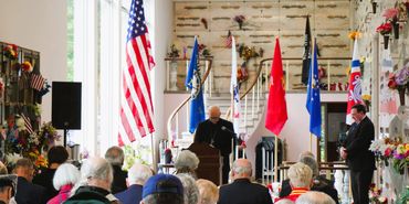 A speaker addresses an audience in a memorial hall with flags and rifles on display.