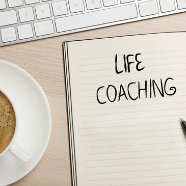 Open notebook with 'LIFE COACHING' written, pen, coffee cup, and keyboard on desk.