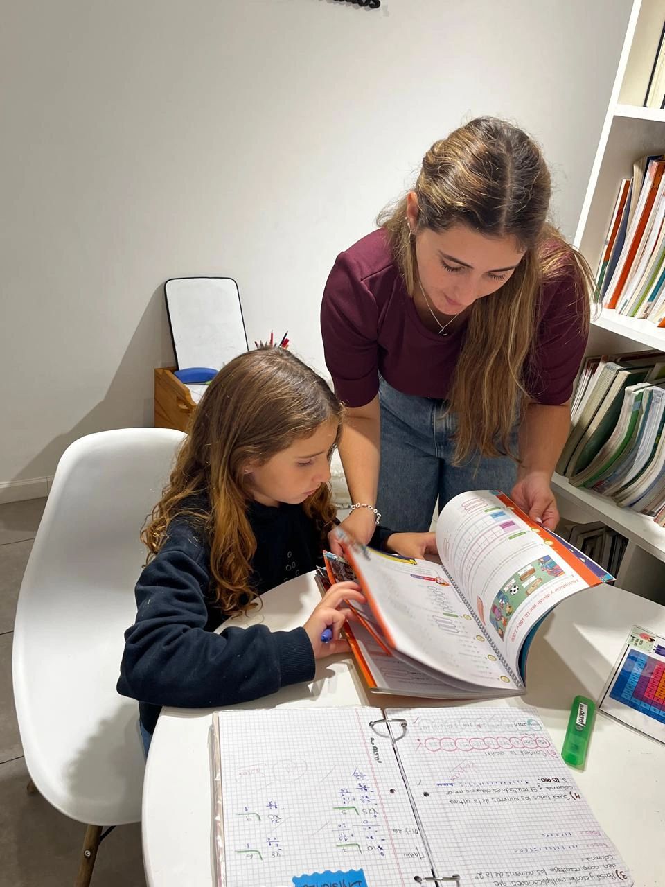 Woman helps girl with homework at a white table filled with notebooks and stationery.