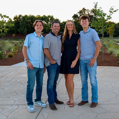 A smiling family of four posing outdoors on a paved area with greenery behind.