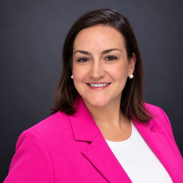 Woman smiling, wearing a bright pink blazer and pearl earrings.