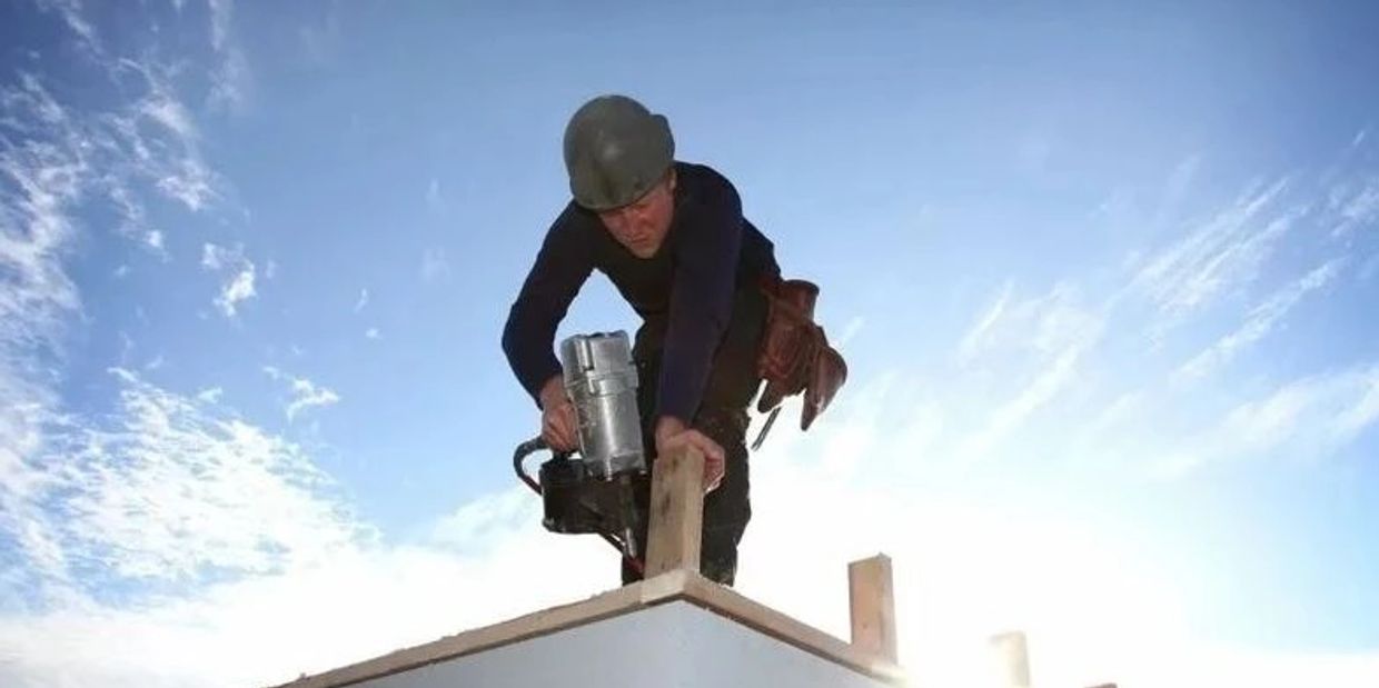 Construction worker using a nail gun on a rooftop under a bright sky.