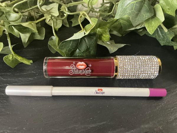 A red lipstick and lip liner from Shawn Marie on a black surface with green leaves.