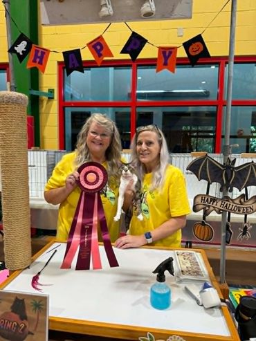 Two women holding a cat and a large ribbon at a Halloween-themed event.