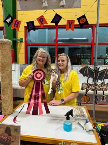 Two women holding a cat and a large ribbon at a Halloween-themed event.