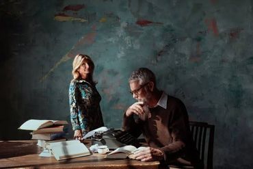 An older man reading a book while a woman stands nearby in a dimly lit room.