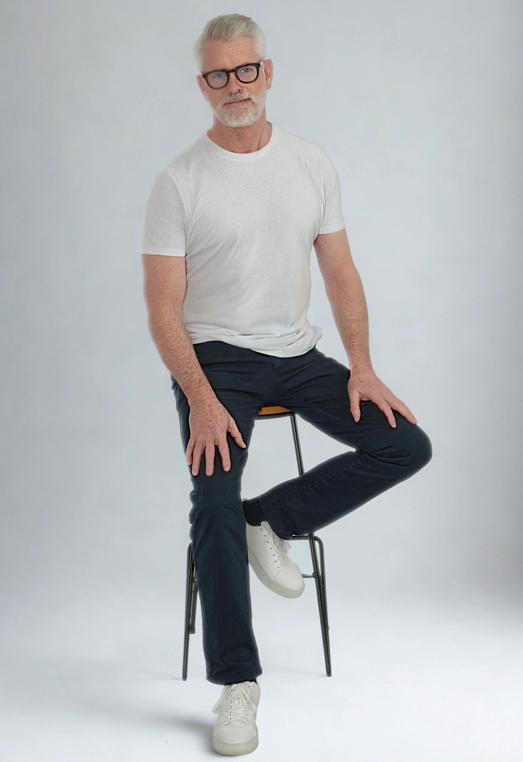 Older man with white hair and glasses sitting on a stool against a plain background.