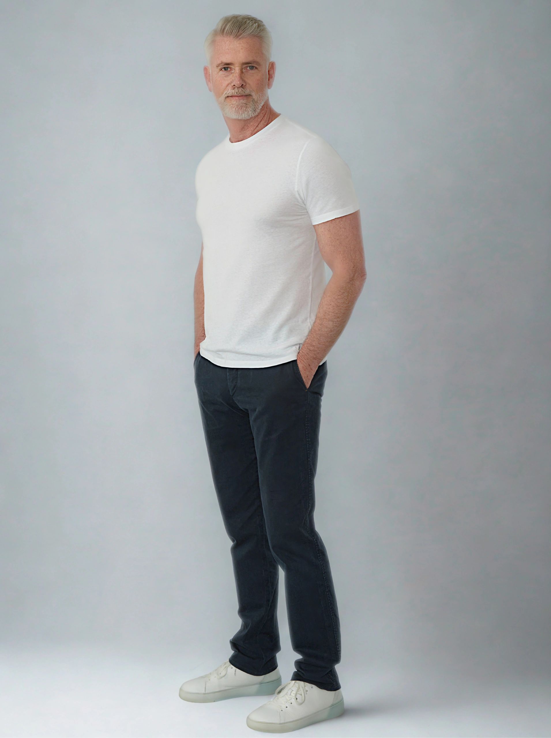 Confident mature man in casual white t-shirt and dark pants against a plain background.