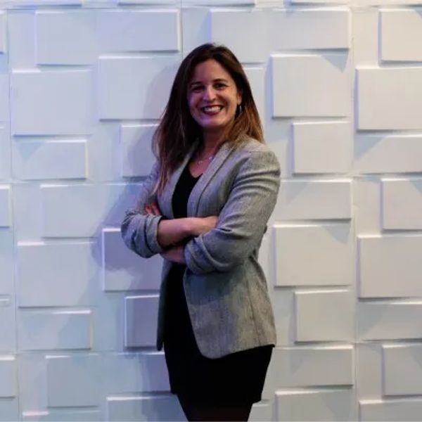 Mujer sonriente con blazer y vestido negro frente a pared blanca texturizada.