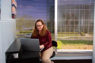 Woman working on laptop by window with blinds in cozy room.