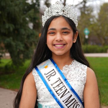 Young girl wearing a crown and Jr. Teen Miss sash smiles outdoors.