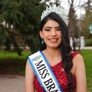 A smiling beauty queen wearing a crown and Miss Brazil sash outdoors.