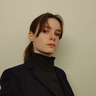 Young woman with brown hair in a ponytail wearing a black turtleneck and blazer.