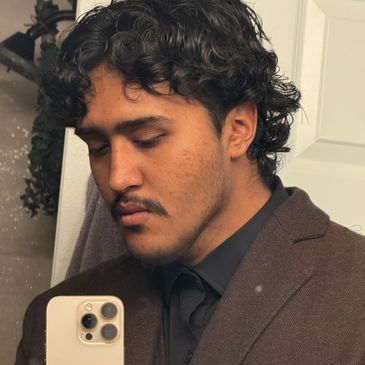 Young man with curly hair in a brown suit taking a selfie.