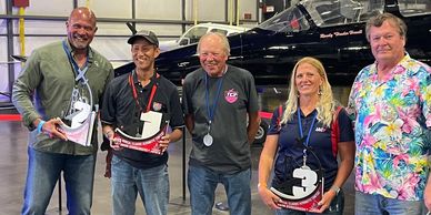 Five adults stand in front of a small airplane holding racing trophies shaped like numbers.
