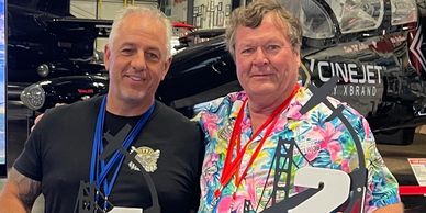 Two men posing with a drone at an indoor event with aircraft in the background.