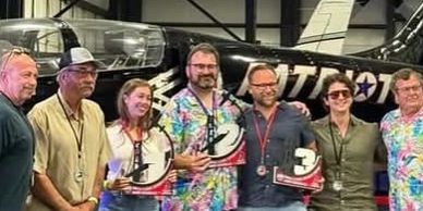 Group of people posing with racing trophies in front of a helicopter.