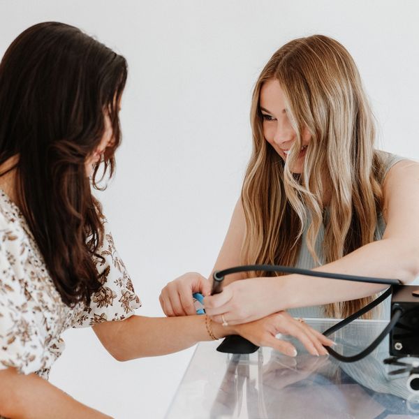 Two young women using a tattoo machine on a wrist at a clear table.