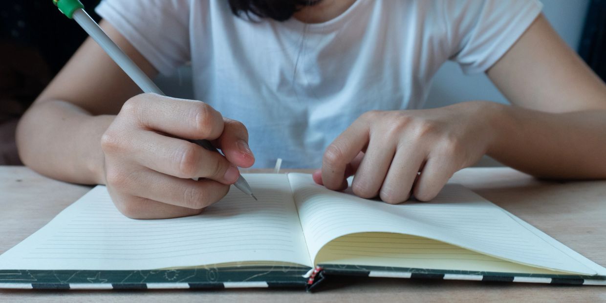 Person writing in a notebook with a pen at a wooden table.