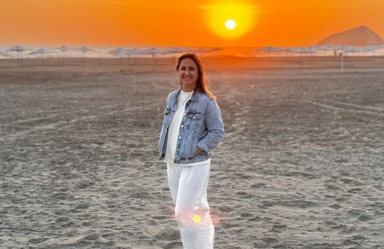 Woman smiling on a beach at sunset, wearing a denim jacket and white pants.