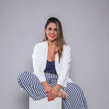 Smiling woman in white blazer and striped pants sitting cross-legged.