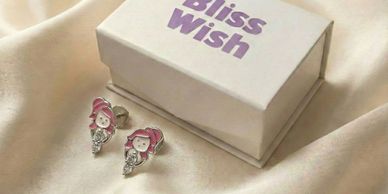 Cute girl-shaped earrings next to a Bliss Wish jewelry box on satin fabric.