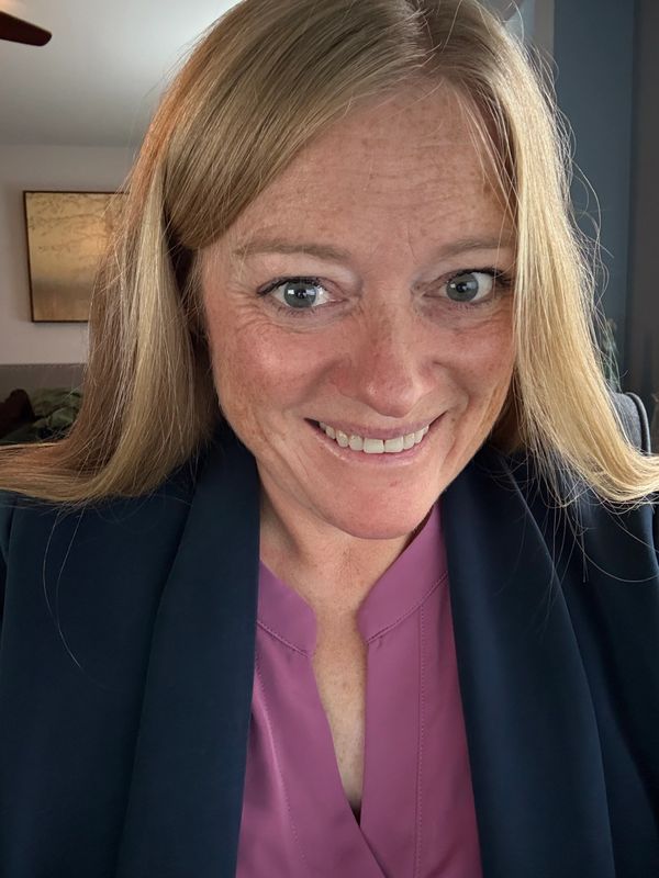 Smiling woman with blonde hair wearing a black jacket and purple top.