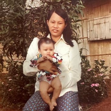 A woman holding a baby outdoors, surrounded by greenery and flowers.