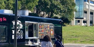Two people ordering food at a black taco food truck outdoors.