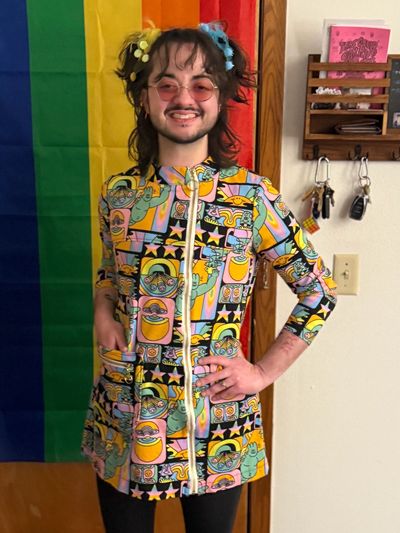 Person in colorful patterned jacket stands in front of rainbow flag, smiling.