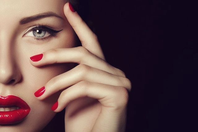 Close-up of a woman's face with red lipstick and nails against a dark background.