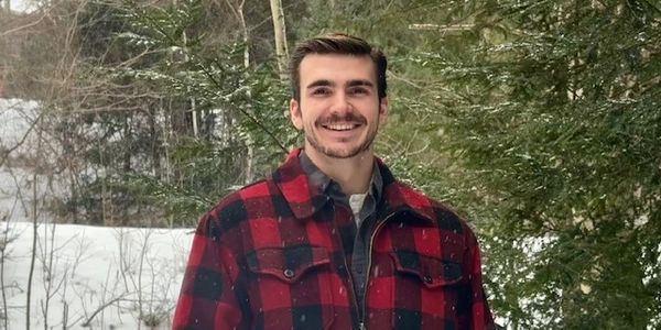 Smiling man in a red and black checkered jacket in a snowy forest.