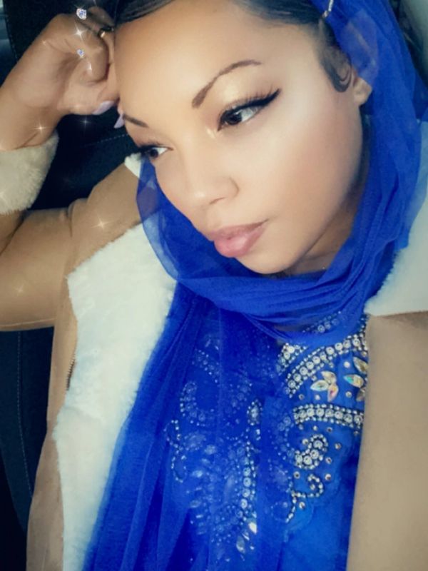 A woman in a blue headscarf and beige coat looks thoughtfully to the side.