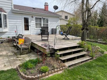 A backyard patio with wooden deck, chairs, steps, and garden beds around.