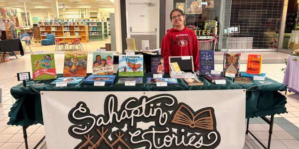 A person stands behind a table showcasing diverse books at a Sahaptin Stories booth.